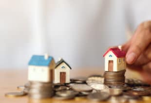 Image d'illustration représentant un tas de pièces sur une table et trois petites maisons en figurine dont l'une est tenue par une main d'homme