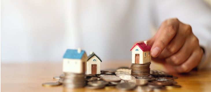 Image d'illustration représentant un tas de pièces sur une table et trois petites maisons en figurine dont l'une est tenue par une main d'homme